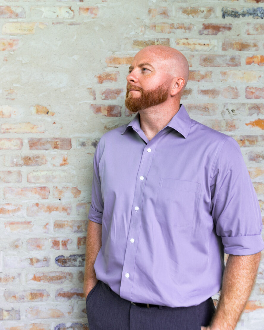 A man with a shaved head and red beard, wearing a light purple dress shirt with rolled-up sleeves, stands against a brick wall, looking to the side with a relaxed expression.
